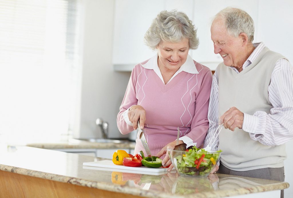 Senior couple in the kitchen smiling and cutting vegetables for a salad.